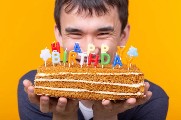 Közeli fel vicces fiatal ázsiai fickó egy sapkát, és egy égő gyertyát, és egy házi torta a kezében pózol a sárga háttér. Évforduló-és születésnapi koncepció. — Stock Fotó