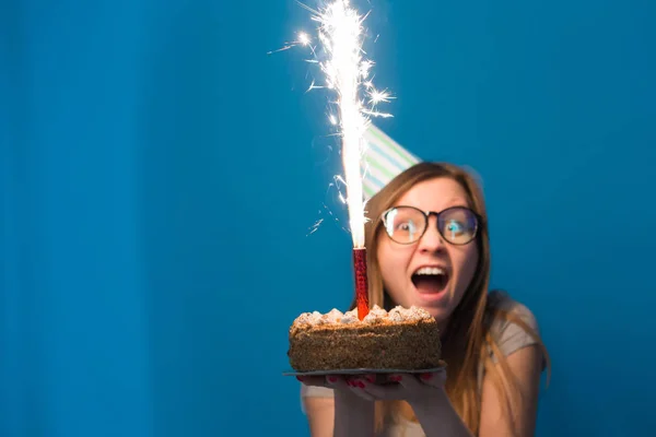 Χαρούμενος νέος θολή κορίτσι μαθητής σε γυαλιά κρατώντας ένα συγχαρητήριο κέικ με ένα κερί που στέκεται σε μπλε φόντο. Ιδέα γενεθλίων. — Φωτογραφία Αρχείου