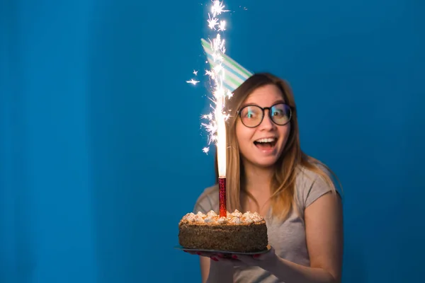 Vidám ifjú homályos lány hallgató szemüveg kezében gratuláló torta egy gyertyát állva a kék háttérben. Születésnapi koncepció. — Stock Fotó