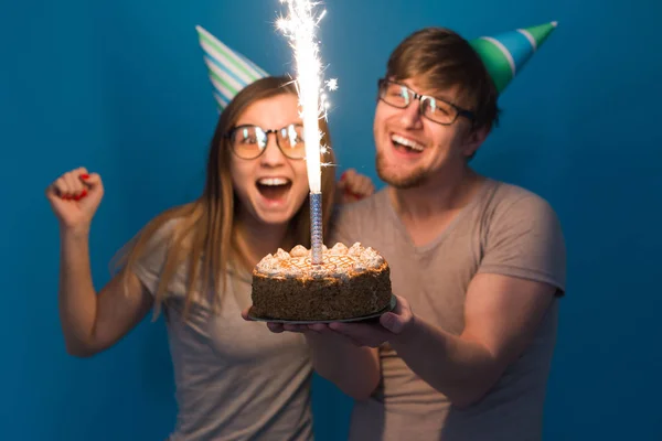 Funny nerd man and woman are wearing holidays caps and glasses holding birthday cake with candles over blue background