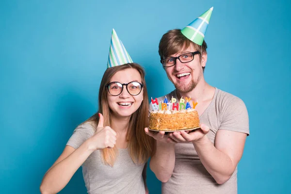 Pareja joven divertida en gorras de papel y con un pastel hacen una cara tonta y desean feliz cumpleaños mientras están de pie sobre un fondo azul. Concepto de felicitaciones y bromas . —  Fotos de Stock