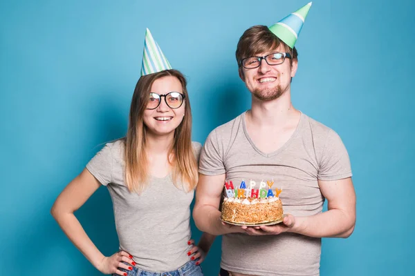 Divertido nerd hombre y mujer están usando días festivos gorras y gafas celebración de pastel de cumpleaños con velas sobre fondo azul —  Fotos de Stock