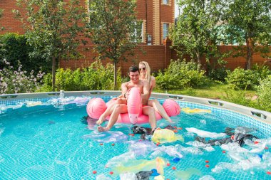 Çöp sorunu, plastik geri dönüşüm, kirlilik ve çevre kavramı - aptal adam ve kadın yüzmek ve kirli bir havuzda eğlenin. Şişeler ve plastik torbalar onlara yakın yüzer