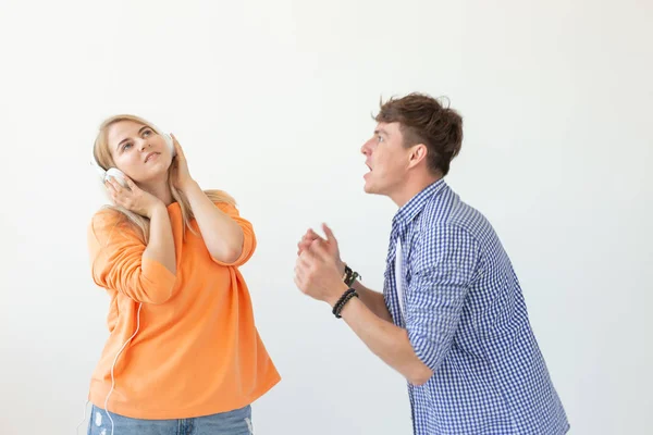 Young upset man begs his woman to listen to him but she listens to music with headphones posing on a white background. Misunderstanding and unwillingness to engage in dialogue. — Stock Photo, Image