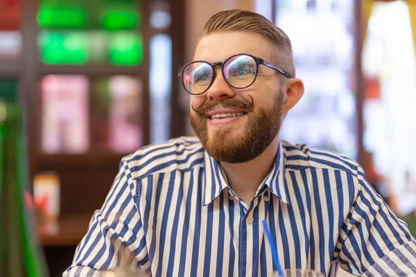 Retrato de un joven guapo con bigote y barba sentado en un café sobre un fondo borroso. El concepto de estudiante o los hombres con estilo . — Foto de Stock