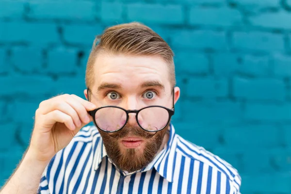Retrato de cerca de un curioso joven sorprendido con gafas con bigote y barba posando sobre un fondo de pared de ladrillo borroso azul. Concepto de sorpresa e información impactante . — Foto de Stock
