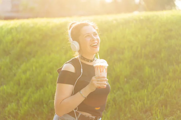 Портрет позитивної молодої красивої дівчини в панк-одязі і окулярах з молочним коктейлем в її руках позує в літньому парку в теплий літній вечір. Концепція відпустки . — стокове фото