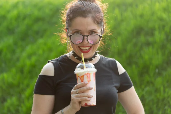 Şık punk giysileri ve gözlük sıcak bir yaz akşamı parkta bir yürüyüş sırasında elinde bir milkshake tutan olumlu neşeli genç kadın. Şehirde rahatlama ve eğlence kavramı.