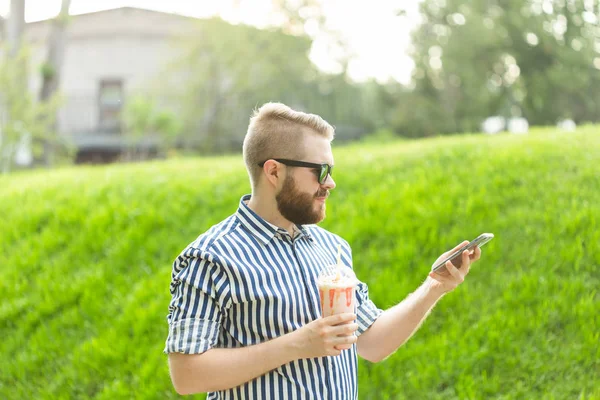 Vista lateral de un joven elegante con una barba sosteniendo un batido y admirando las vistas de la ciudad caminando en el parque en un cálido día de verano. El concepto de descanso y relajación . — Foto de Stock