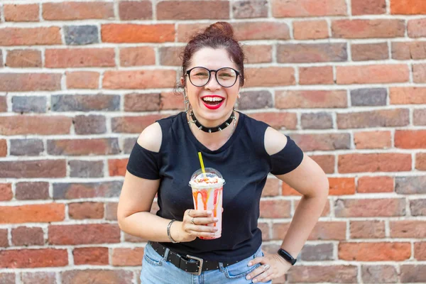 Şık giysiler içinde Sevimli neşeli genç kadın gülüyor ve bir tuğla duvara karşı elinde bir milkshake tutarak. Şehirde bir yaz tatili kavramı. Copyspace.