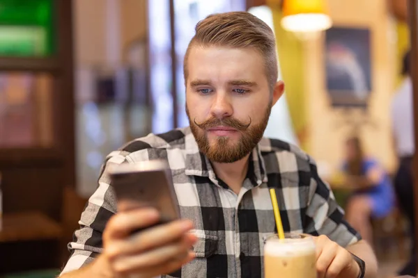 Un joven elegante con bigote y barba está viendo una red social usando un teléfono inteligente mientras está sentado en un café un fin de semana. El concepto de dependencia de las redes sociales . — Foto de Stock