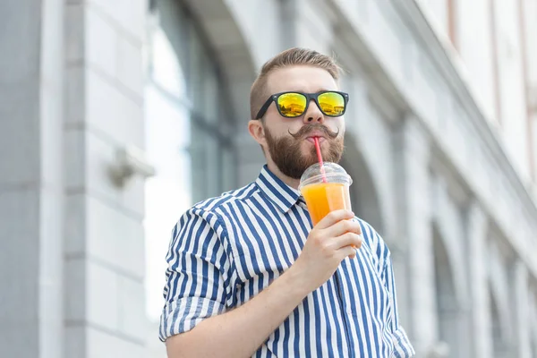 Retrato de un joven hipster borroso y elegante con barba y bigote sosteniendo jugo de naranja fresco en sus manos. Concepto de snack saludable . — Foto de Stock