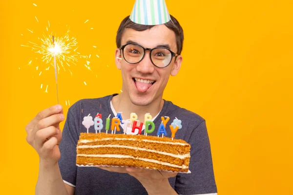 Lustiger positiver Typ mit Brille hält eine selbstgebackene Torte mit der Aufschrift "Happy Birthday" in den Händen, die auf gelbem Hintergrund posiert. Konzept von Feiertagen und Jahrestagen. — Stockfoto