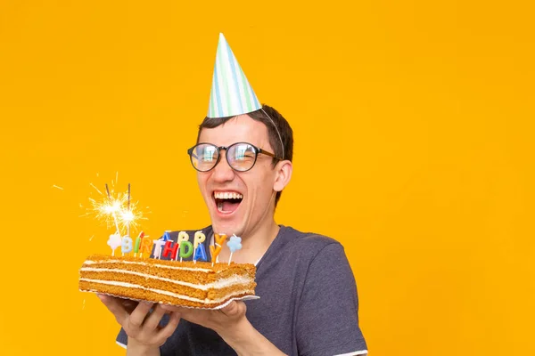 Porträtt av en rolig positiv kille med ett papper mössa och glasögon innehar en gratulationer hemlagad tårta i händerna på en gul bakgrund med kopia utrymme. Koncept och nöje och firande. — Stockfoto