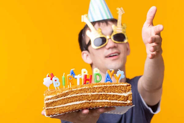 Portrait of a funny positive guy with a paper cap and glasses holding a congratulatory homemade cake in his hands on a yellow background. Concept and fun and celebration. — Stock Photo, Image