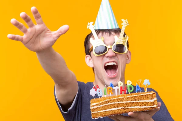 Positif drôle jeune asiatique gars avec une casquette et une bougie brûlante et un gâteau fait maison dans ses mains posant sur un fond jaune. Concept anniversaire et anniversaire . — Photo
