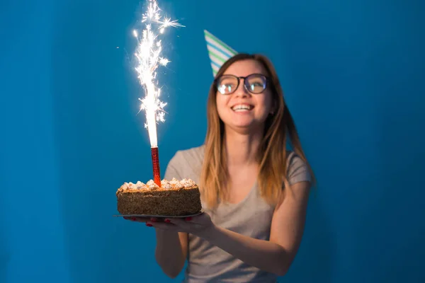 Jovem estudante menina borrada alegre em óculos segurando um bolo de parabéns com uma vela em pé sobre um fundo azul. Conceito de aniversário . — Fotografia de Stock