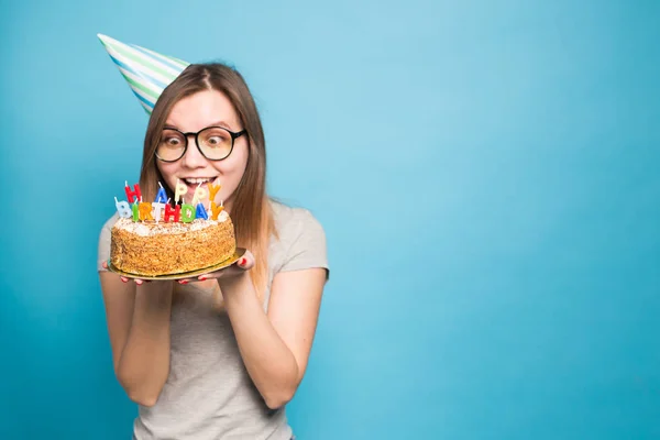 Szalona zabawna dziewczyna w papierowym kapeluszu i okularach posiadających duży tort urodzinowy na niebieskim tle z przestrzenią kopiową. Koncepcja Prank i pozdrowienia. — Zdjęcie stockowe