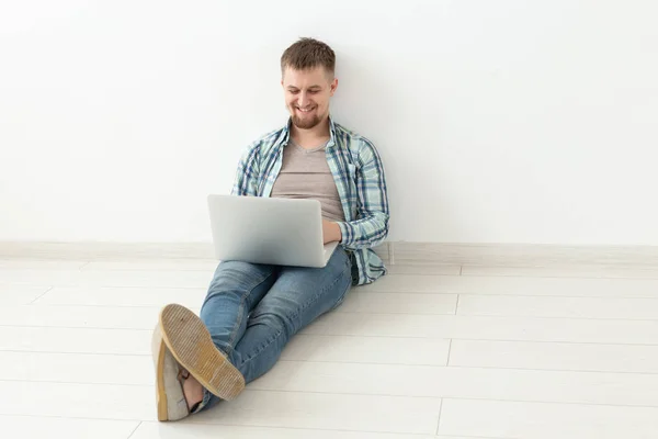Pozytywny młody człowiek w casual ubrania surfowanie po Internecie w poszukiwaniu nowych mieszkań siedzi na podłodze w pustym pomieszczeniu. Koncepcja znalezienia mieszkania za pomocą Internetu i laptopa. — Zdjęcie stockowe
