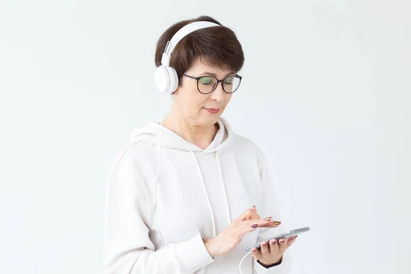 Cute positive middle-aged woman in sweater and glasses is listening to music with wire headphones standing on a white background and holding tablet . Concept of hobbies and subscriptions to favorite — Stock Photo, Image