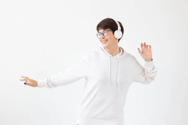 Cute positive middle-aged woman in sweater and glasses is listening to music with wire headphones standing on a white background. Concept of hobbies and subscriptions to favorite radio station. — Stock Photo, Image
