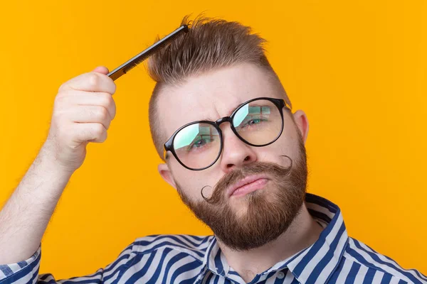 Retrato de un joven hipster guapo divertido con bigote y barba peinándose el pelo sobre un fondo amarillo. Concepto de barbería y autocuidado . — Foto de Stock
