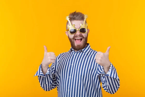 Joven hipster alegre levantando sus pulgares en gafas con guitarras posando sobre un fondo amarillo. El concepto de una fiesta y unas vacaciones exitosas — Foto de Stock