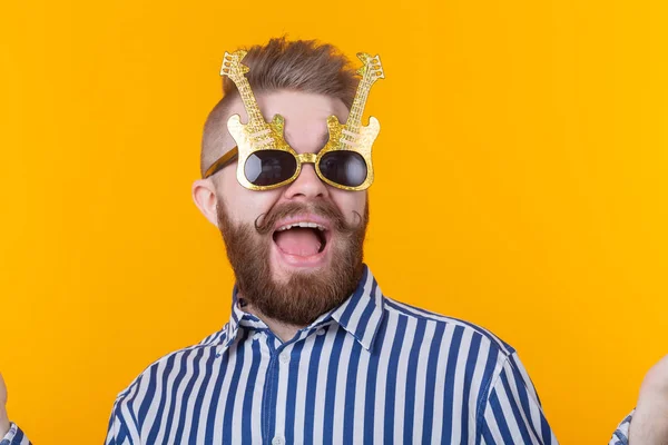 Joven positivo con gafas en forma de guitarras se regocija sobre un fondo amarillo. El concepto de celebración y fiestas . — Foto de Stock