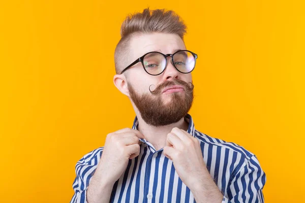 Lindo joven estudiante hipster con un bigote y una barba con gafas endereza su camisa mientras posa sobre un fondo amarillo. El concepto de confianza en sí mismo . — Foto de Stock