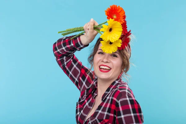 격자 무늬 셔츠와 파란색 배경 위에 포즈를 취하는 아름다운 밝은 거베라 꽃을 스니핑 붕대에서 아름다운 긍정적 인 젊은 여성. 선물과 인사의 개념. — 스톡 사진