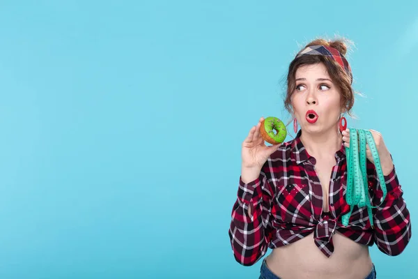 Close-up shocked surprised young beautiful woman looking at measuring tape and holding a green donut in hands posing on a blue background copyspace. Concept of diet and rejection of harmful high