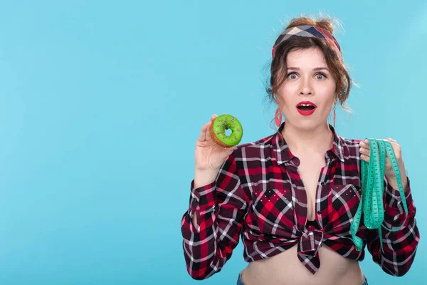 Close-up shocked surprised young beautiful woman looking at measuring tape and holding a green donut in hands posing on a blue background with copy space. Concept of diet and rejection of harmful high