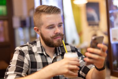 Bıyıklı ve sakallı şık genç adam, hafta sonu bir kafede otururken akıllı telefon kullanarak bir sosyal ağ izliyor. Sosyal ağlara bağımlılık kavramı.