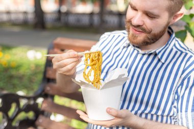 Güneşli bir yaz gününde bir park bankında otururken bir öğle yemeği kutusundan Çin erişte yiyen genç yakışıklı hipster adam Yan görünümü. Sağlıklı ve besleyici bir atıştırmalık kavramı.