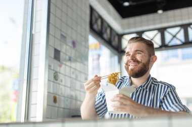 Yakışıklı genç hipster adam bir kafede oturan ve pencereye bakarak ahşap çubukları kullanarak Çin erişte yeme. Asya mutfağı konsepti.