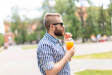 Sıcak güneşli bir yaz gününde parkta yürürken bir saman ile meyve suyu bir bardak ile olumlu neşeli bir genç adam Portresi. Çalışma sonrası dinlenme kavramı ve hafta sonları çalışma.