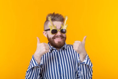 Neşeli hipster genç adam sarı bir arka plan aleyhine poz gitar ile gözlük başparmak yukarı yükselterek. Başarılı bir parti ve tatil kavramı