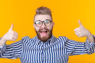 Bir bıyık ve sakal ile neşeli yakışıklı genç hipster adam başparmak gösteren sarı bir arka plan üzerinde poz. Telefon ve iletişim konsepti.