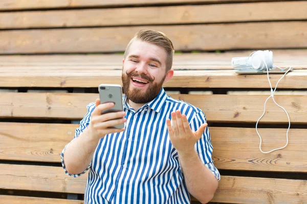 Joven estudiante positivo con bigote y barba hablando con sus amigos en video comunicación usando smartphone y wi-fi gratis Internet sentado en el parque en un banco con una tableta y auriculares . — Foto de Stock