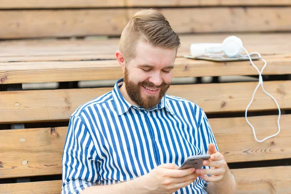 Pensativo joven estudiante hipster masculino serio con un bigote y barba escribiendo sms mensaje a sus amigos descansando después de estudiar en el parque sentado en un banco de madera con una tableta y auriculares — Foto de Stock