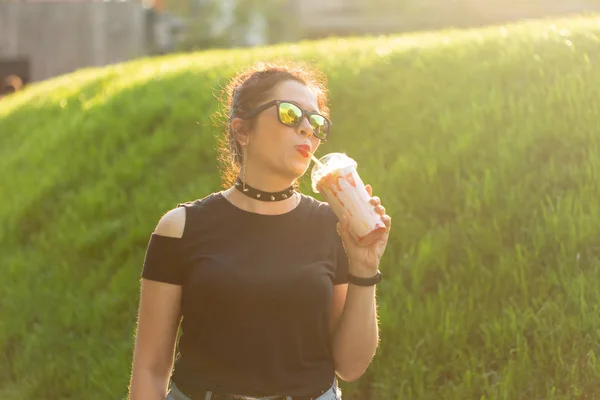 Portret pozytywnej młodej dziewczyny w punkowych ubraniach i okularach z Milkshake w rękach stwarzających w letnim parku w ciepłym letnim wieczorem. Koncepcja urlopów. — Zdjęcie stockowe