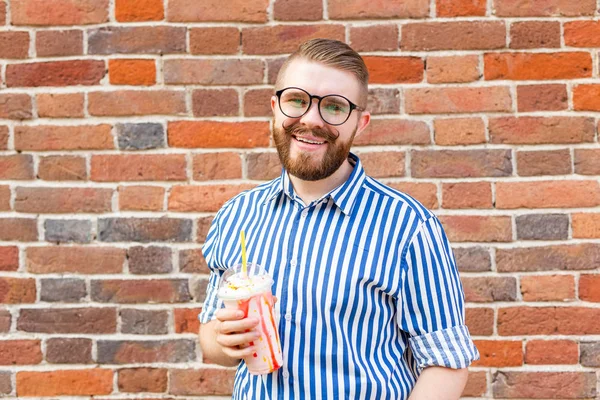 Positive young hipster guy in glasses and stylish beard is drinking a milkshake with a straw while standing outside against a brick wall. The concept of lovers of light sweet drinks.
