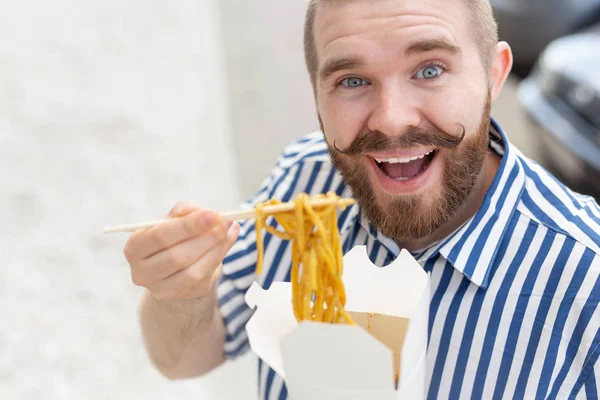 Close-up of an amusing young hipster guy eating chinese noodles with wooden chopsticks sitting in a park outside on a warm summer day. The concept of rest and snacking on the street. — Stock Photo, Image