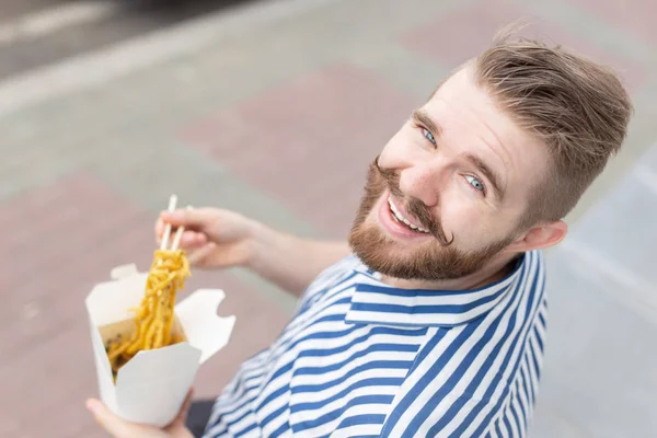 Mladý mužský hipírek s knírkem a vousy konzumuje čínské nudle s dřevěnými hůlky z bílého oběšního boxu. Koncepce asijských kuchyní. — Stock fotografie