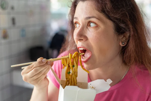 Porträt eines jungen, fröhlichen Mädchens, das in einem Café chinesische Nudeln isst und aus dem Fenster schaut. das Konzept der gesunden asiatischen Küche. — Stockfoto