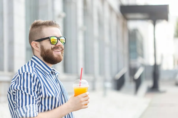 Vista lateral de un joven empresario bastante alegre con un bigote elegante y una barba con jugo en las manos, caminando por la ciudad después de un día de trabajo. Concepto de positivo y descanso . — Foto de Stock