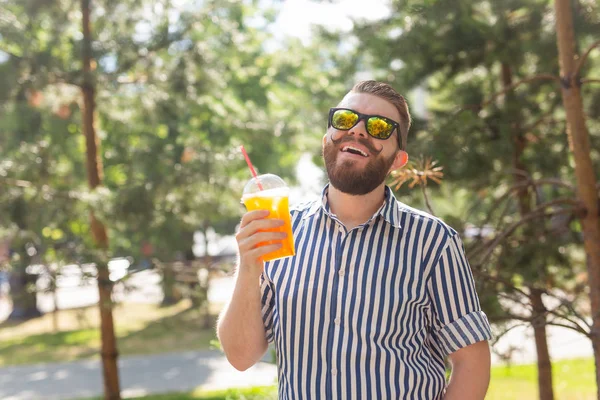 Portret van een positieve vrolijke jongeman met een glas sap met een rietje tijdens het wandelen in het Park op een warme zonnige zomerdag. Het concept van rust na studie en werk in het weekend. — Stockfoto