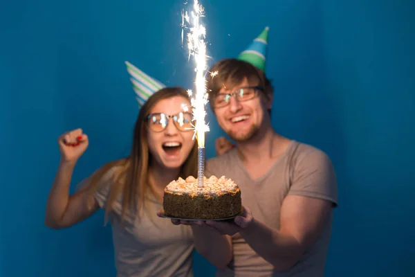 재미 괴상한 남자와 여자는 파란색 배경위에 스파클러와 생일 케이크를 들고 휴일 모자와 안경을 착용 — 스톡 사진