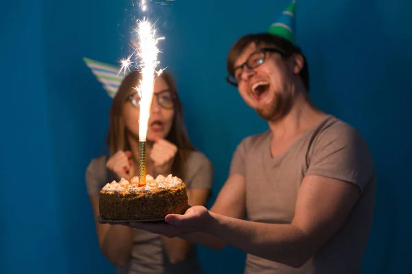 Engraçado nerd homem e mulher estão usando feriados bonés e óculos segurando bolo de aniversário com velas sobre fundo azul — Fotografia de Stock