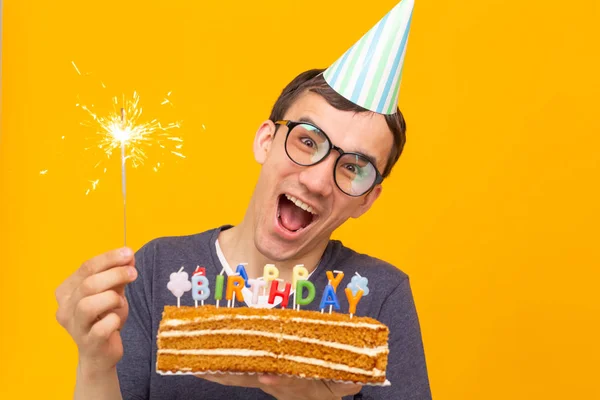 Pazzo allegro giovane uomo in occhiali e carta cappelli di congratulazione in possesso di torte buon compleanno in piedi su uno sfondo giallo. Concetto di congratulazioni giubilari . — Foto Stock
